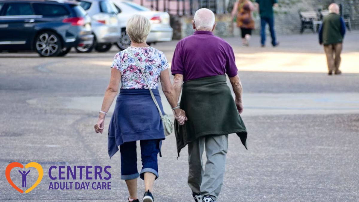 An elderly couple walks hand in hand down the street, their backs to the camera, as cars pass by on the side.