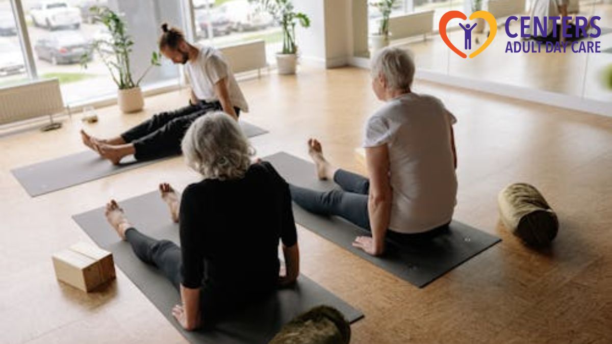 Adult day care services session where two seniors rest on yoga mats between exercise sessions, enjoying the view from a window in an indoor gym.