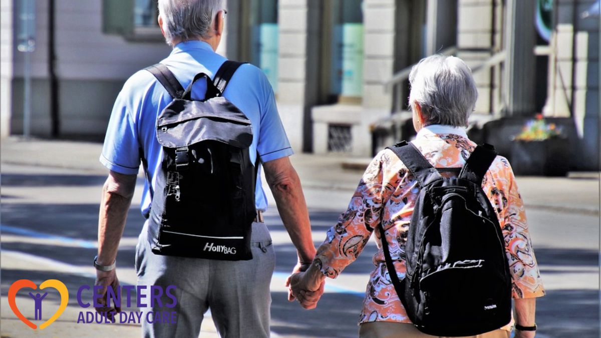 Two seniors from an adult day care center walk hand-in-hand down a sunny afternoon street, their backs to the camera, enjoying a leisurely stroll together.
