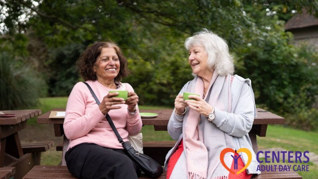 Two women from an adult day care center sit in a sunny, green park, sharing conversation and a warm drink together.