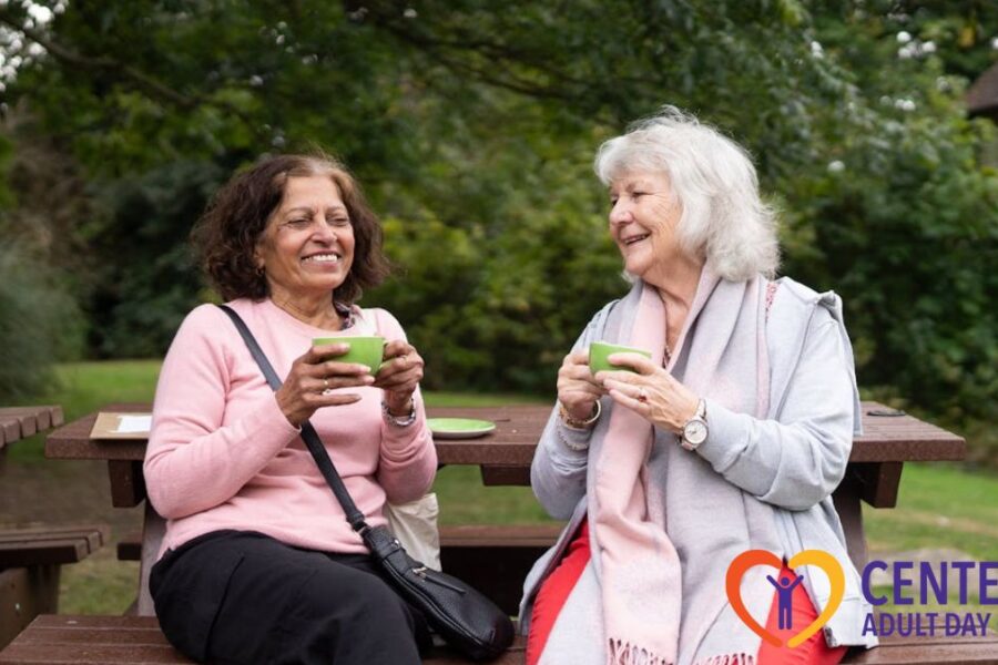 Two women from an adult day care center sit in a sunny, green park, sharing conversation and a warm drink together.