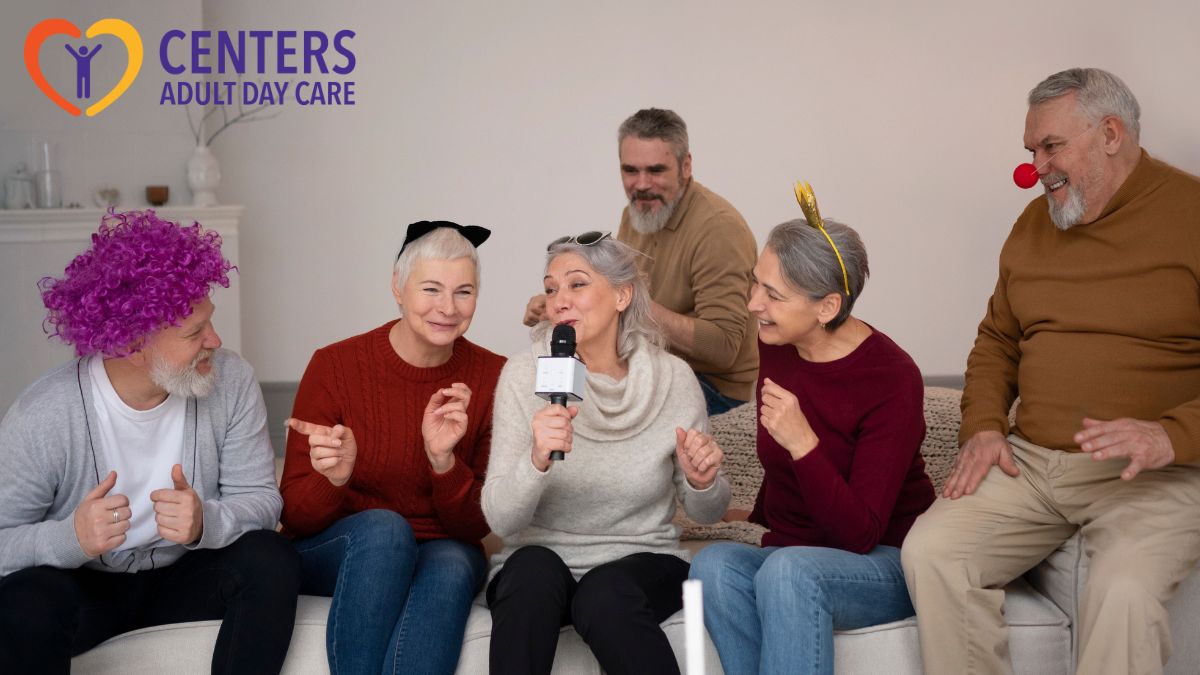 A group of seniors sits on a sofa during an adult day care activity, with a senior woman at the center holding a microphone and speaking to the group.