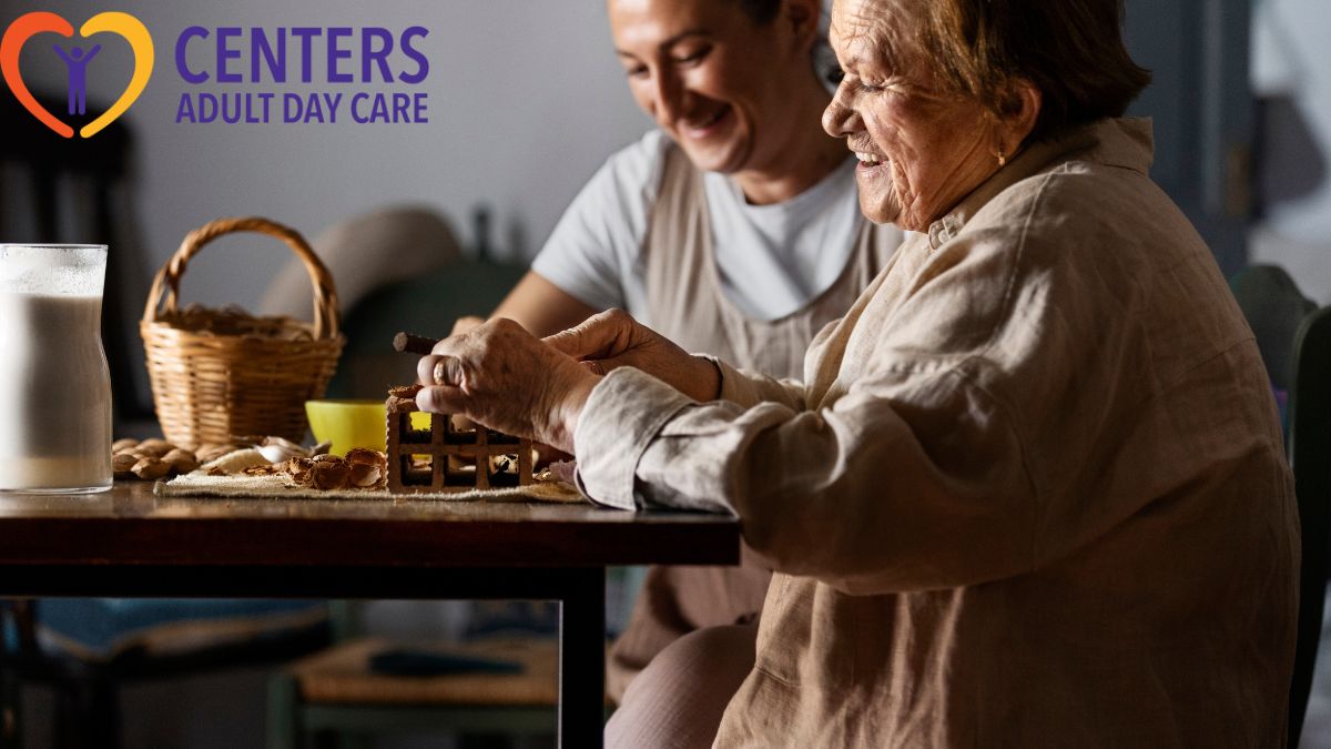 Senior woman receiving guidance from a caregiver on an arts and crafts project, sitting at a large table filled with materials in a bright activity room..