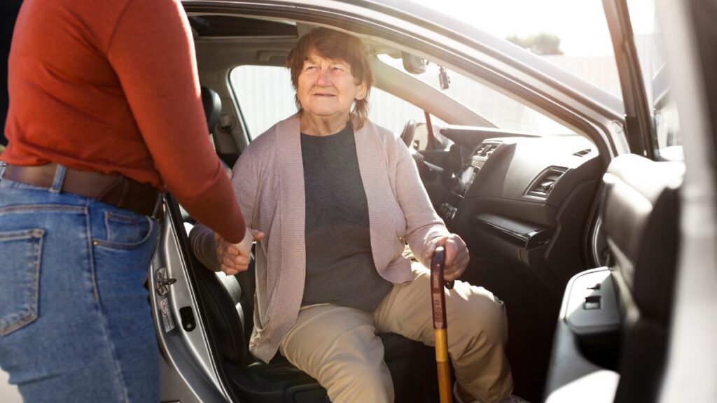 A senior woman holding a cane emerges from a car, receiving gentle assistance to help her stand, illustrating supportive transportation for seniors.