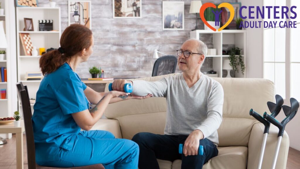 A senior man seated in a chair holds a handheld weight in front of him, guided by his therapist in a brightly lit therapy room.
