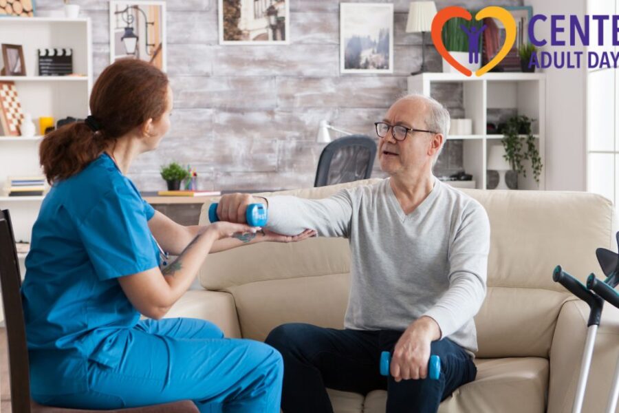 A senior man seated in a chair holds a handheld weight in front of him, guided by his therapist in a brightly lit therapy room.